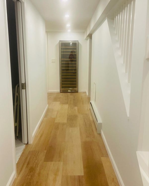 Narrow hallway with wood flooring, white walls, wine cooler, and stairs.
