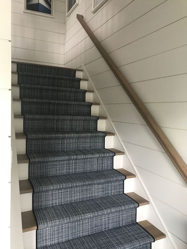 Staircase with gray woven carpet runner, white risers, and a wooden handrail, with white horizontal shiplap walls.