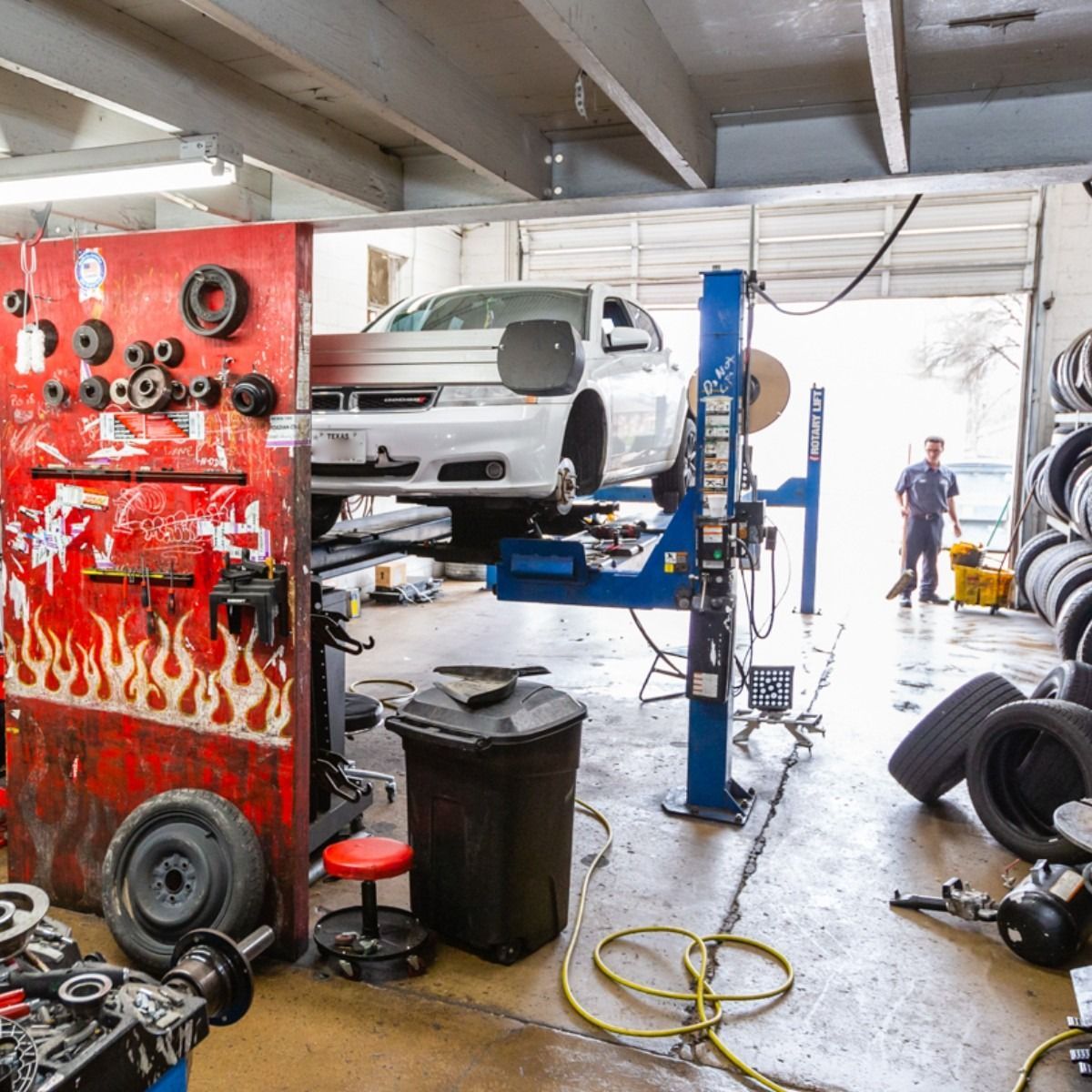 Car on lift in auto repair shop; mechanic in doorway; tools, tires, and equipment around.