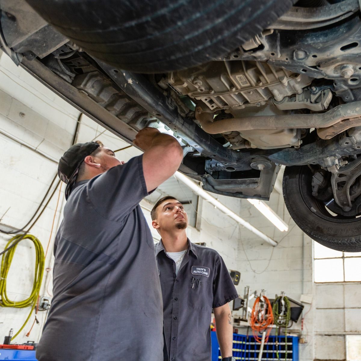 Two mechanics inspect underside of a car in a garage, one using a flashlight.