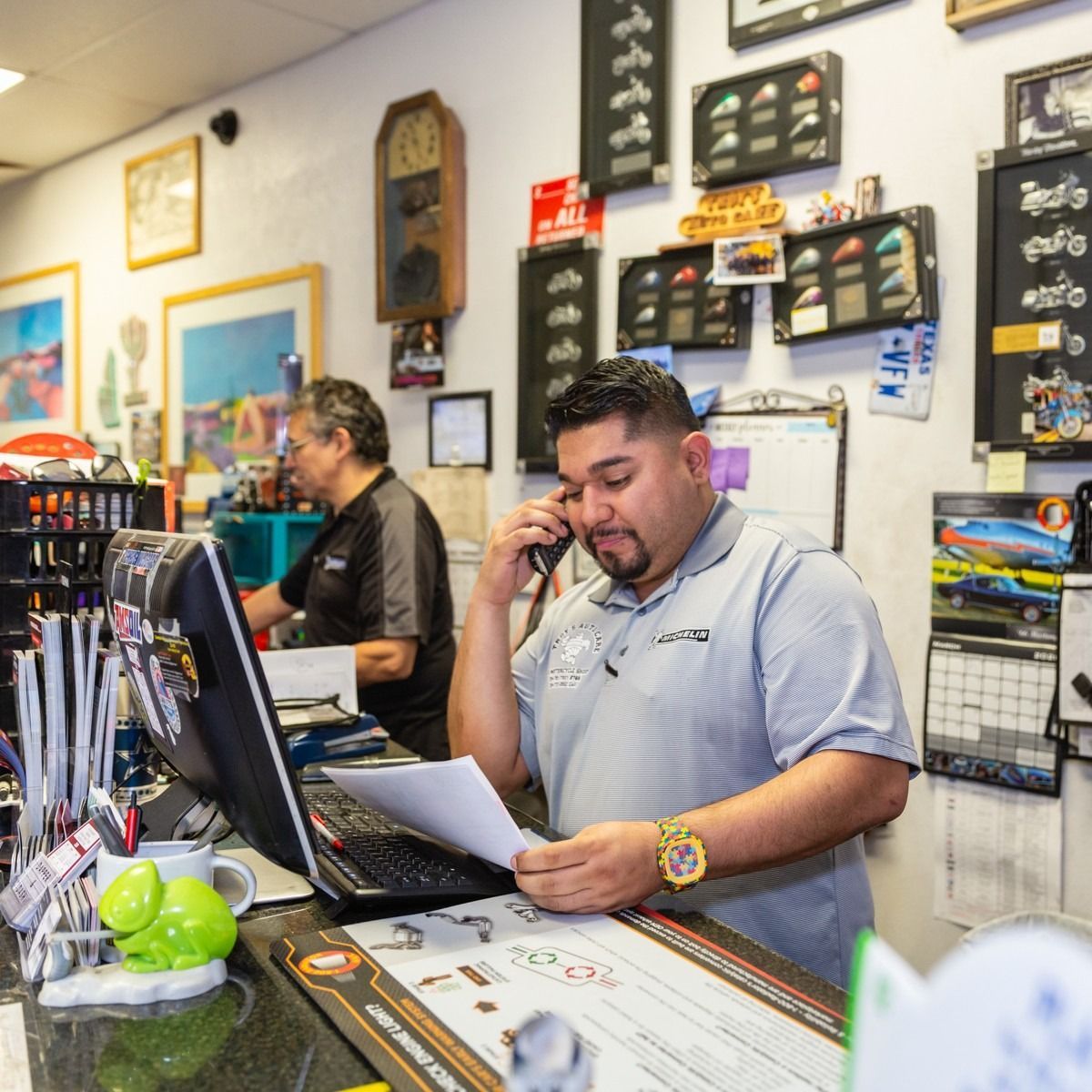 Man on phone at a counter in shop, looking at paperwork. Another person in background.
