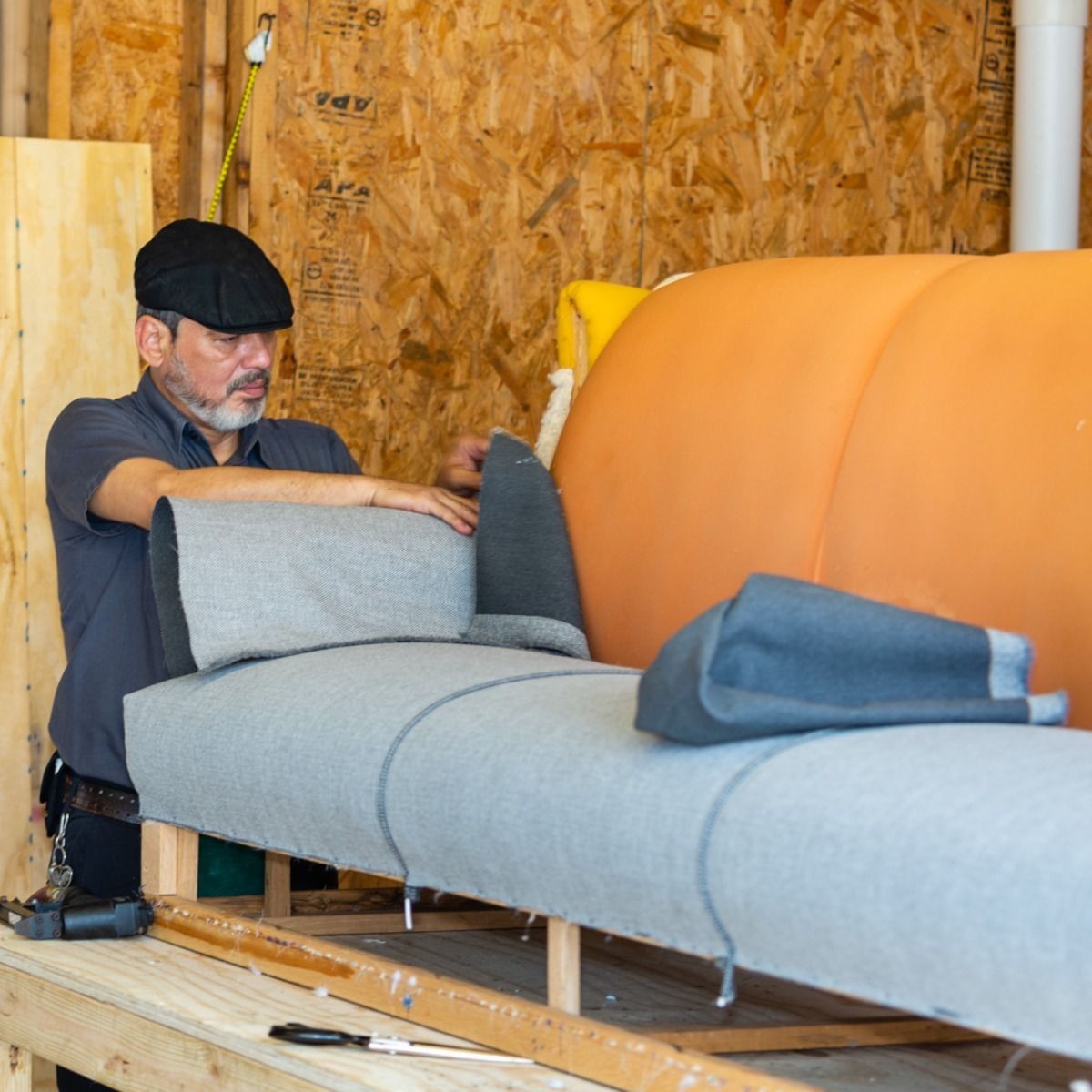 Man assembling a light grey sofa in a workshop, working with fabric. Orange tank in the background.