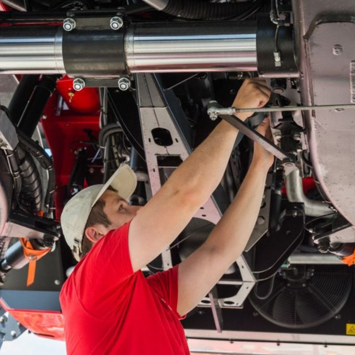 Mechanic in red shirt and hat repairs machinery underneath, using a tool.