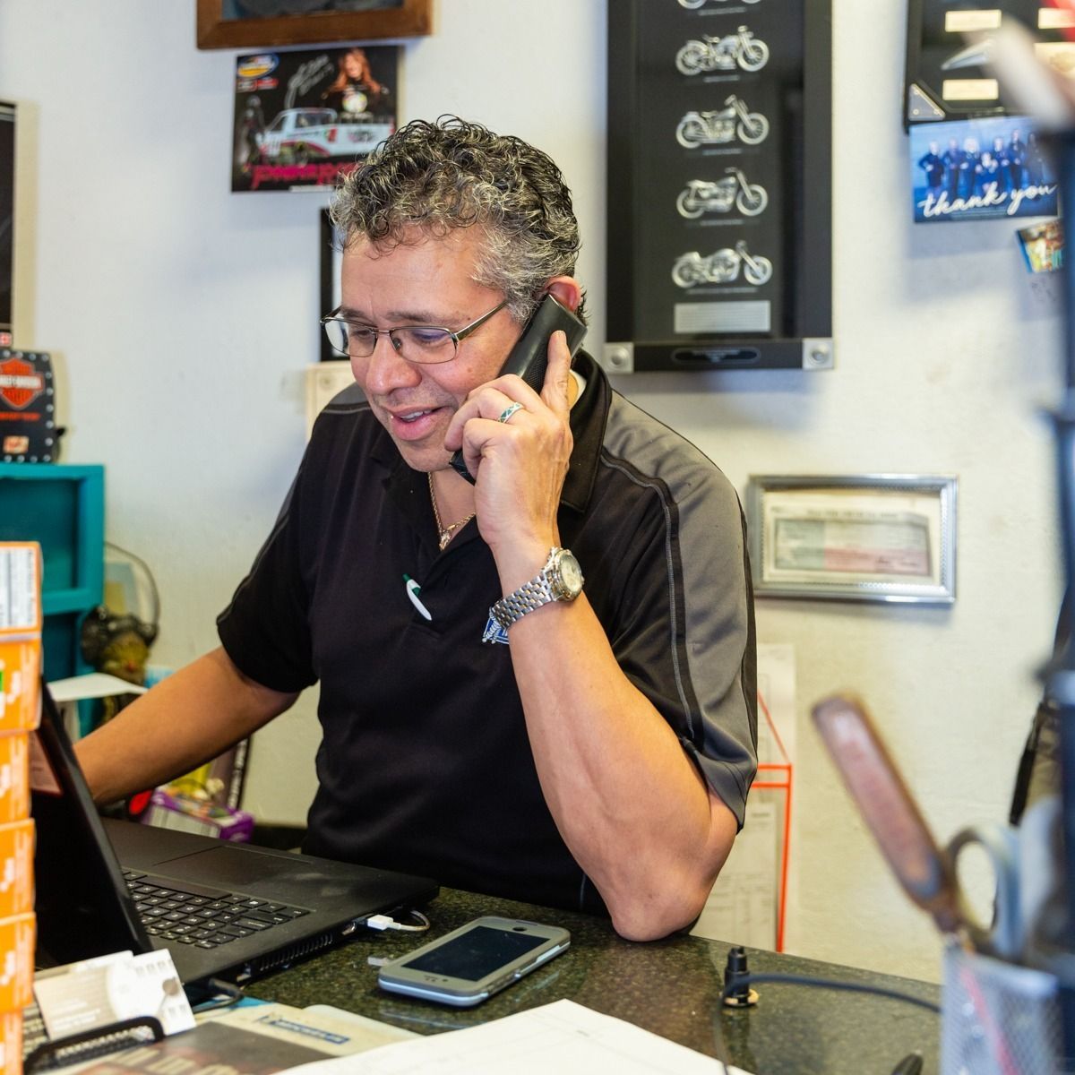 Man on phone at a desk with laptop, inside a shop; motorcycle art on wall.
