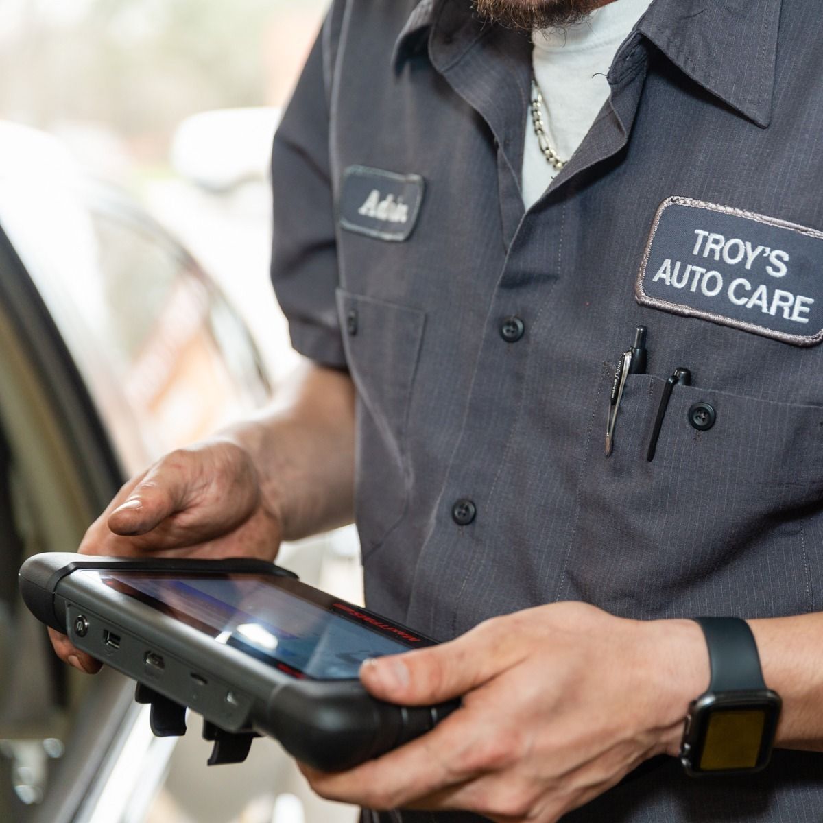 Mechanic in gray uniform holding a diagnostic tablet, working at Troy's Auto Care.