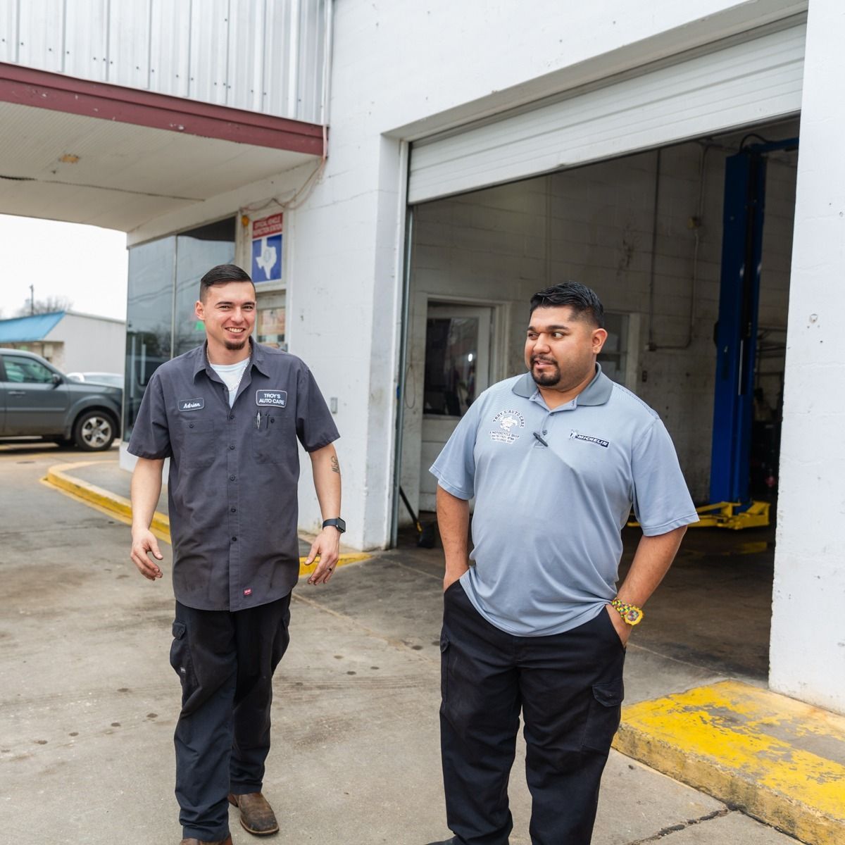 Two men in work attire stand outside a garage. One smiles.