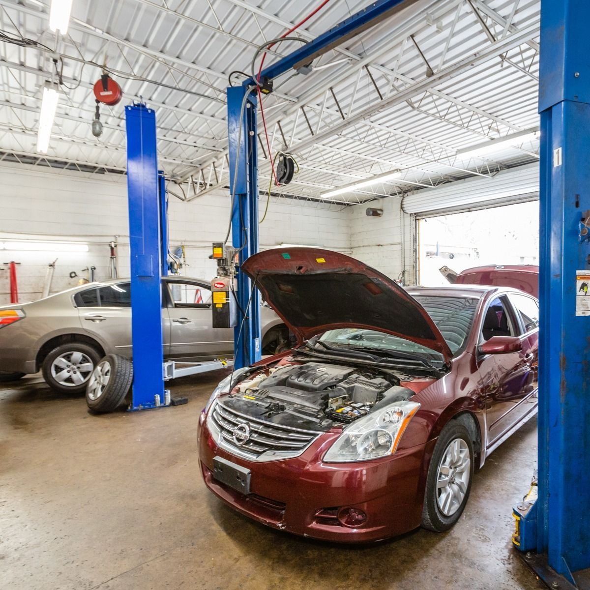 A red car with an open hood in a garage, next to a car raised on a lift.