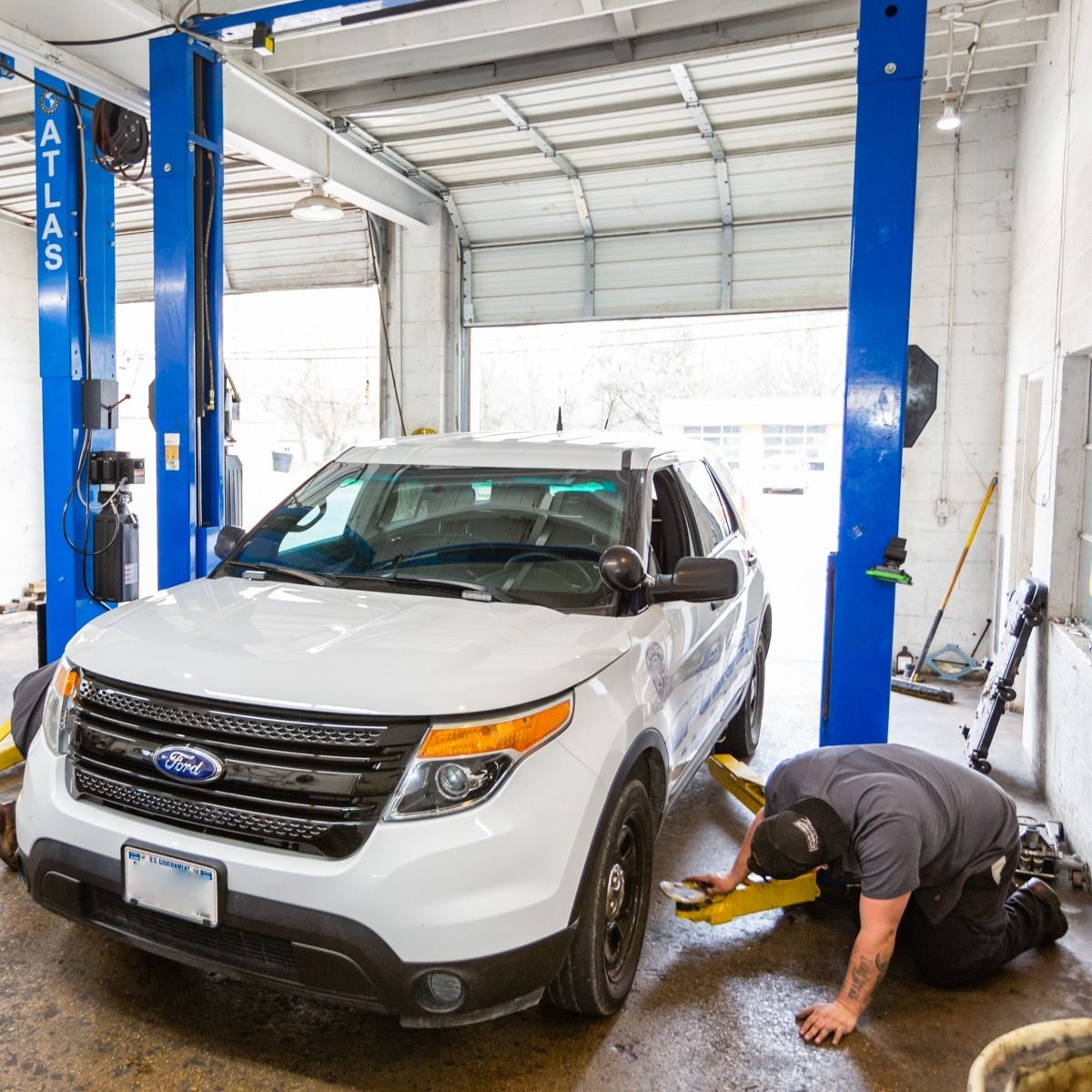 White Ford Explorer police SUV on a lift in a garage, a mechanic kneeling to inspect it.