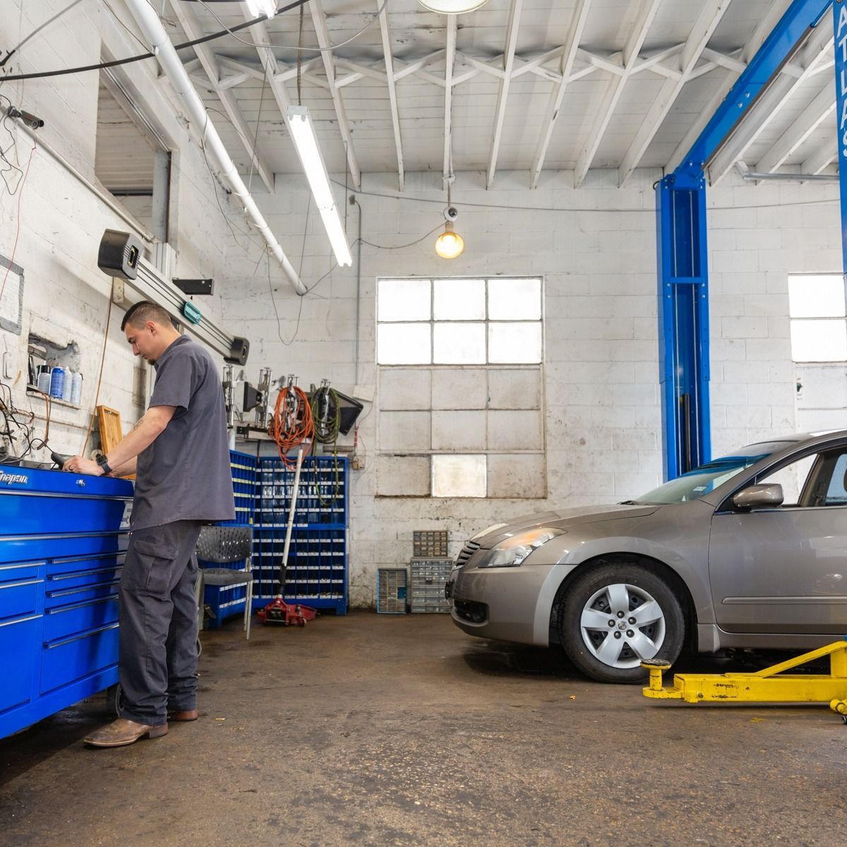 Mechanic working on car in a garage with tools and lift. Grey sedan parked.