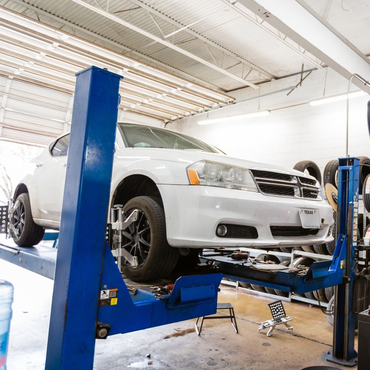 White car on a blue car lift inside a garage, tires stacked in background.