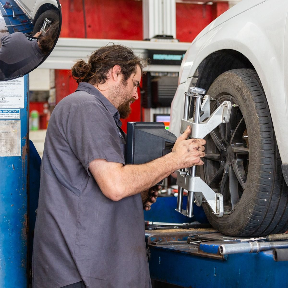 Mechanic adjusting wheel alignment on a white car using diagnostic equipment inside a garage.