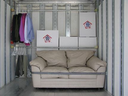 Storage unit with a beige couch, moving boxes, and hanging clothes on metal shelving.