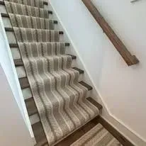 Staircase with striped carpet runner and wooden handrail against a white wall.