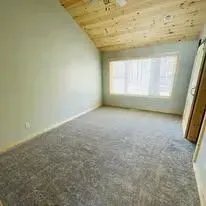 Empty room with gray carpet, angled ceiling, and large window. Wooden door visible.