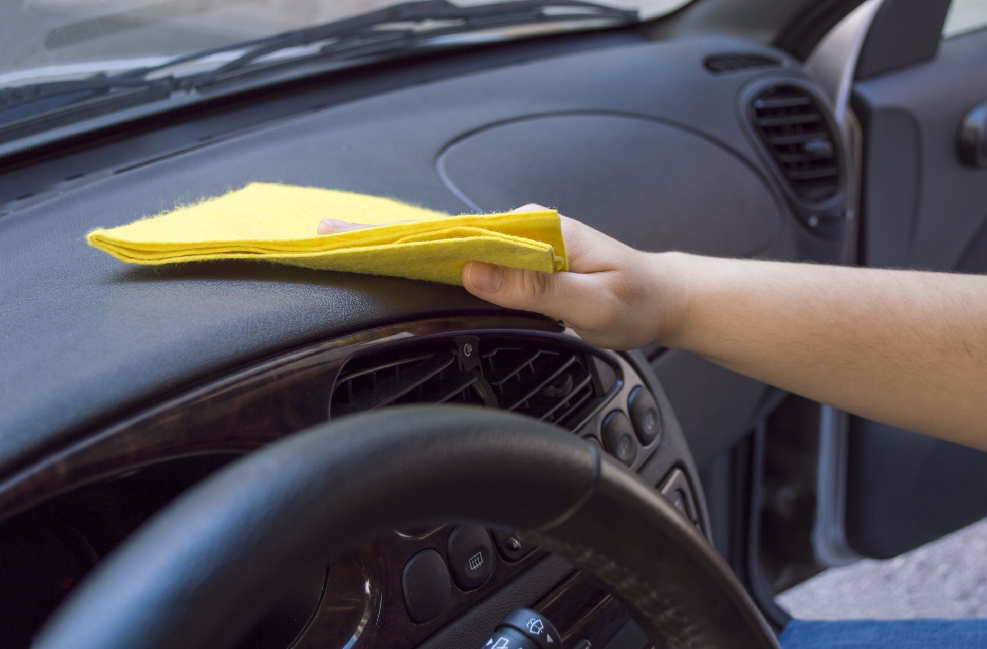 Hand wiping a yellow cloth on a car dashboard, near steering wheel.