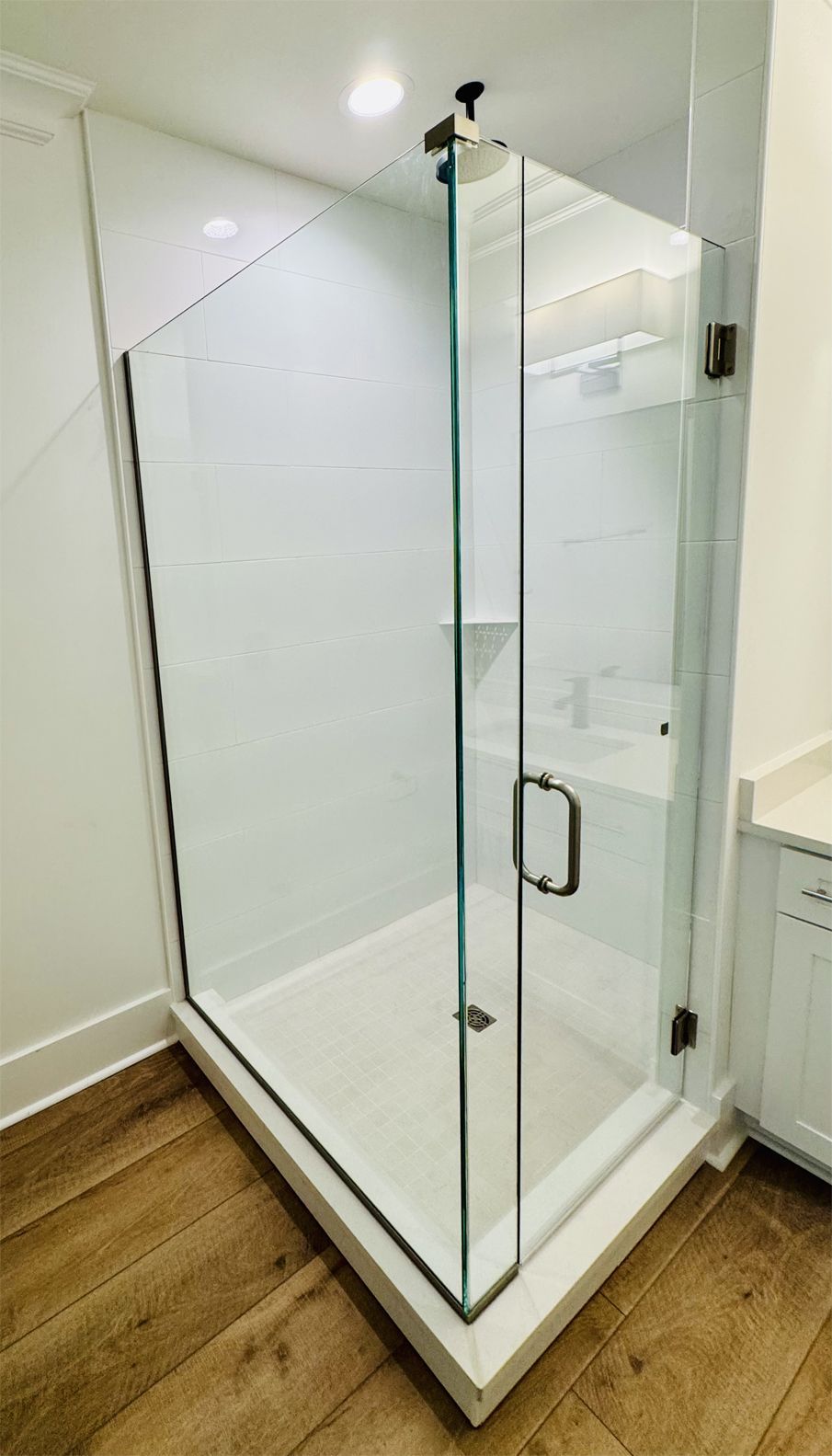 Modern glass shower enclosure in a white-tiled bathroom with light brown wooden flooring.