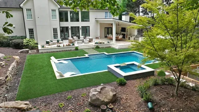 Backyard with pool, patio, and modern house; green lawn, blue water, beige and white house exterior.
