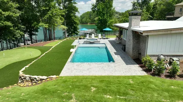 Backyard with pool, putting green, lake view, and a house with a stone chimney on a sunny day.