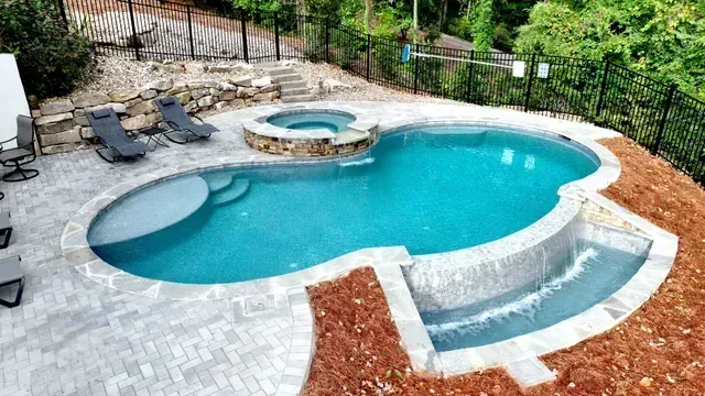 Pool with waterfall and hot tub on a hillside, surrounded by a gray brick patio and black fence.