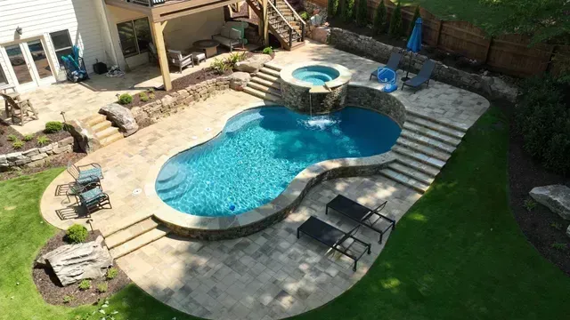 A luxury backyard with a turquoise pool, hot tub, and stone patio, surrounded by green grass.