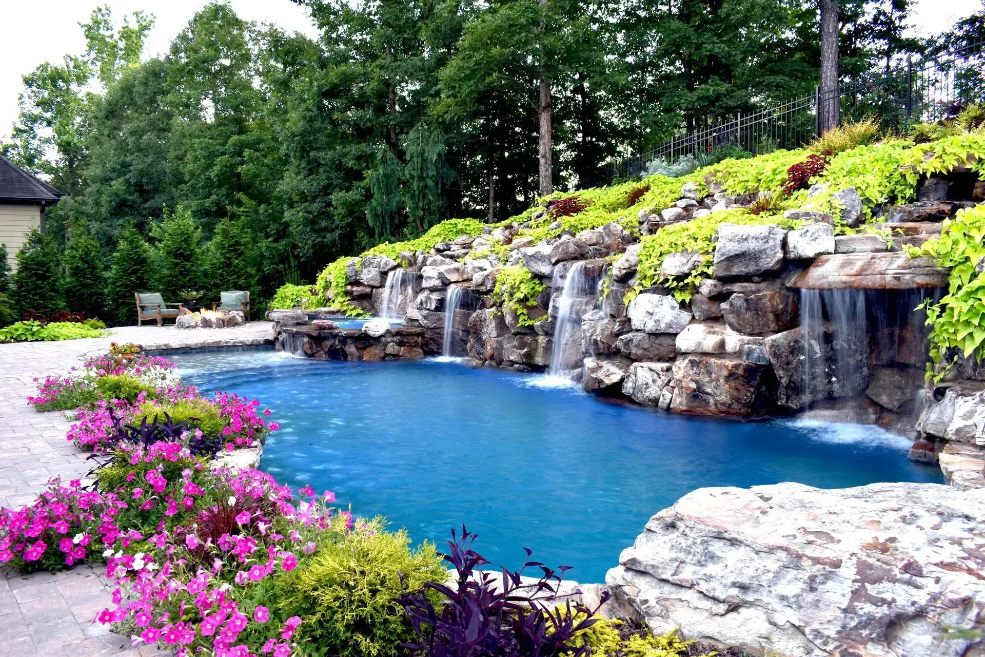 A blue swimming pool with cascading waterfalls, surrounded by rocks, flowers, and greenery.