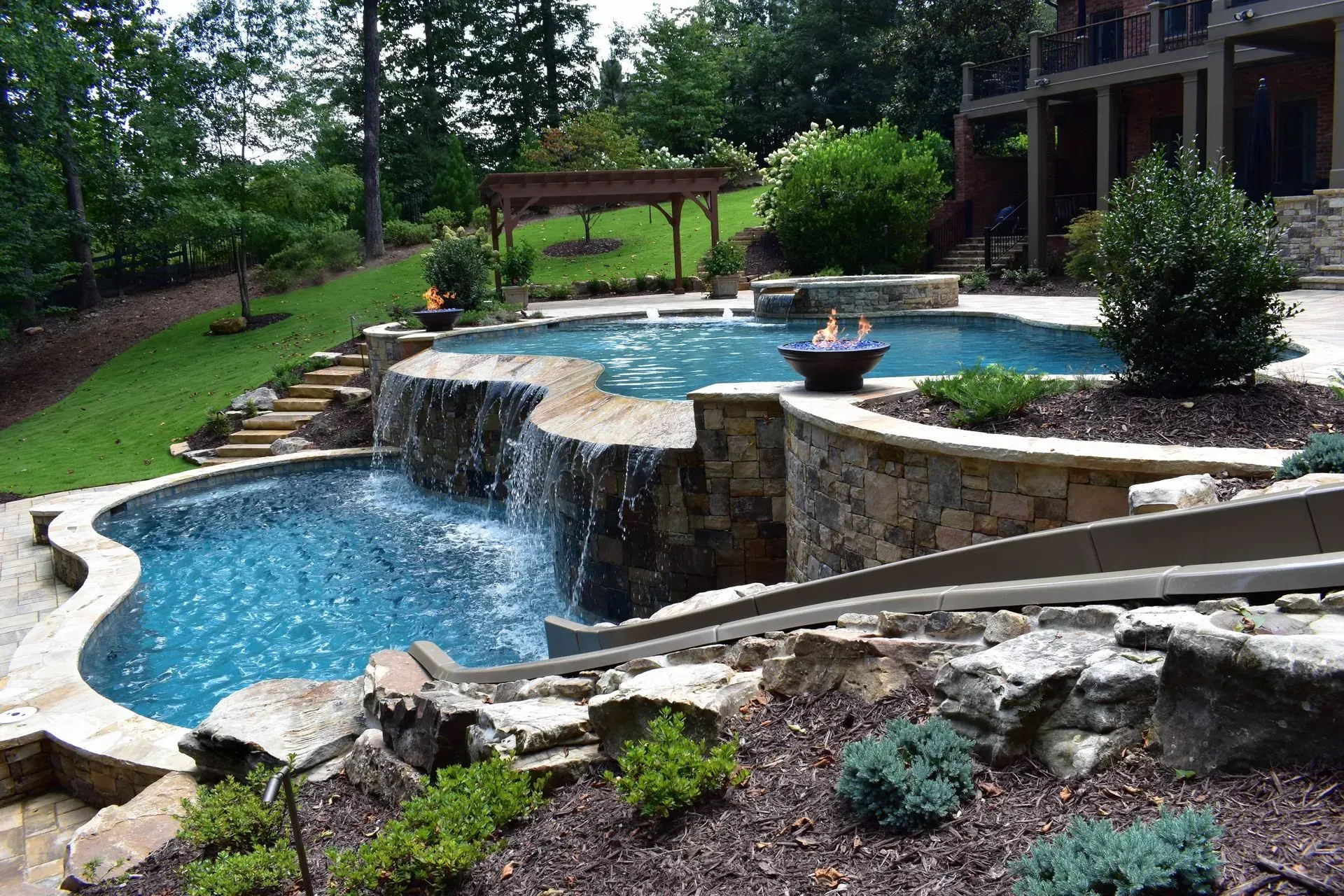 Backyard pool with waterfall, slide, and fire pit surrounded by landscaping and stone walls.