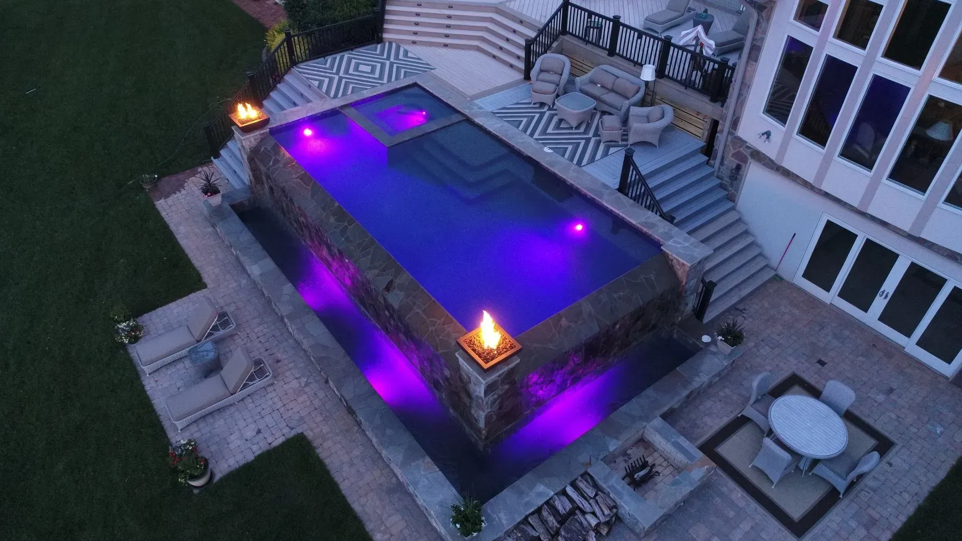 Night view of a luxurious rectangular pool with purple lights, a stone facade, and outdoor seating.