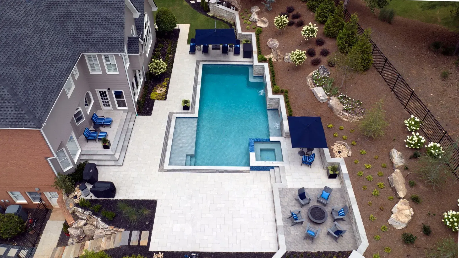 Aerial view of a backyard with a pool, patio, and landscaped areas. The pool is surrounded by light paving stones and dark blue accents.