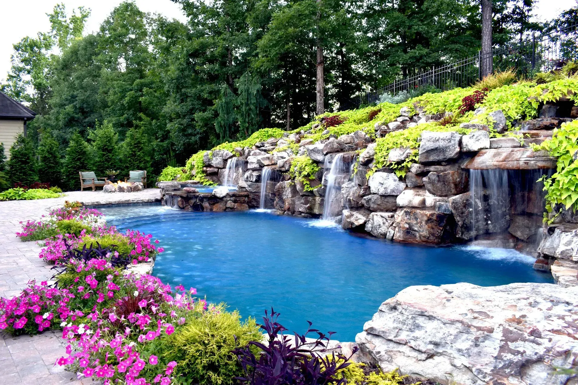 Swimming pool with waterfall feature, surrounded by rocks, flowers, and lush greenery.
