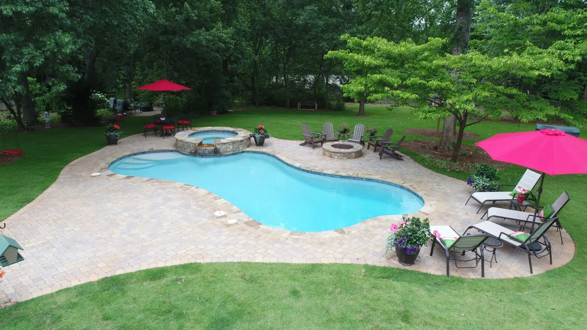 Backyard pool with spa, lounge chairs, red umbrellas, and a fire pit surrounded by landscaping.
