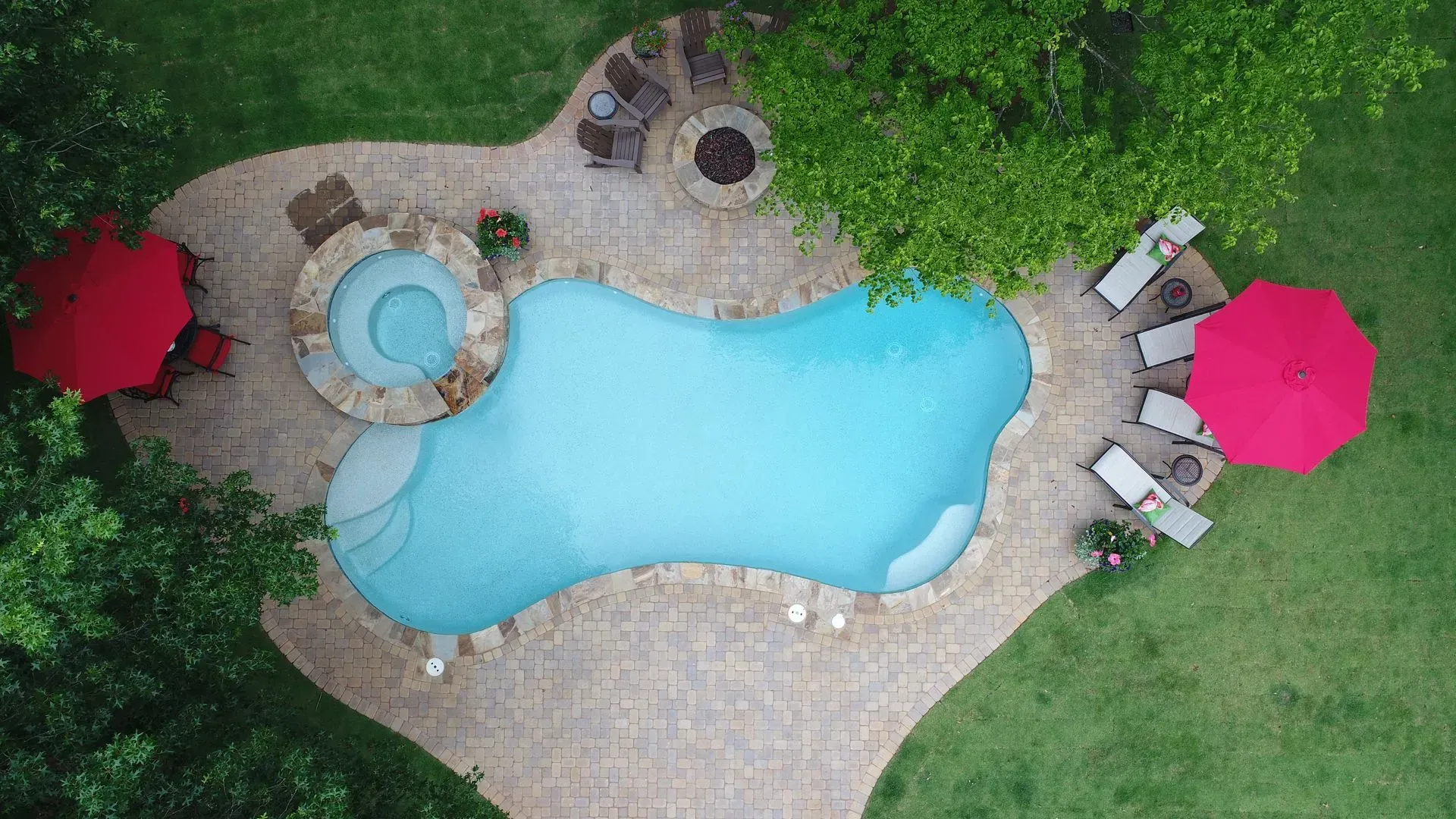 Overhead view of a pool with a hot tub, lounge chairs, and red umbrellas on a stone patio surrounded by grass and trees.