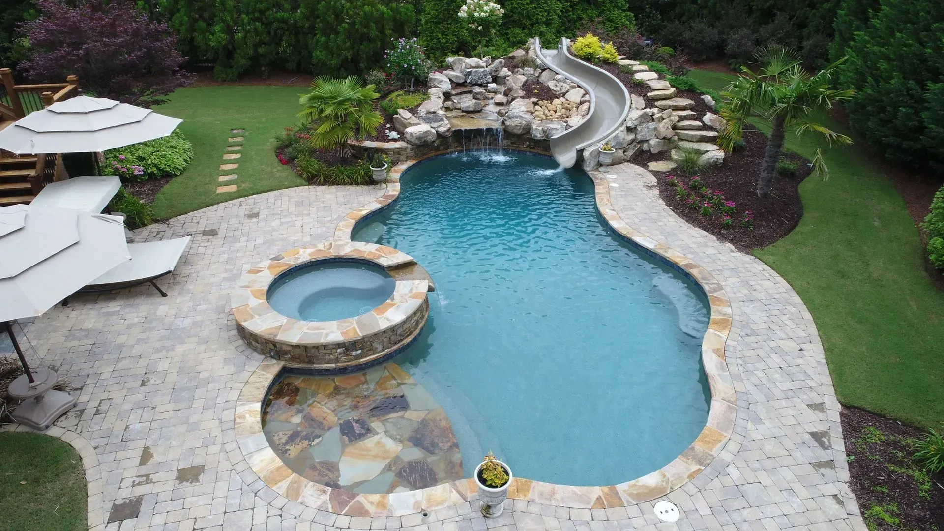 Swimming pool with a waterslide, hot tub, and stone patio surrounded by grass and landscaping.