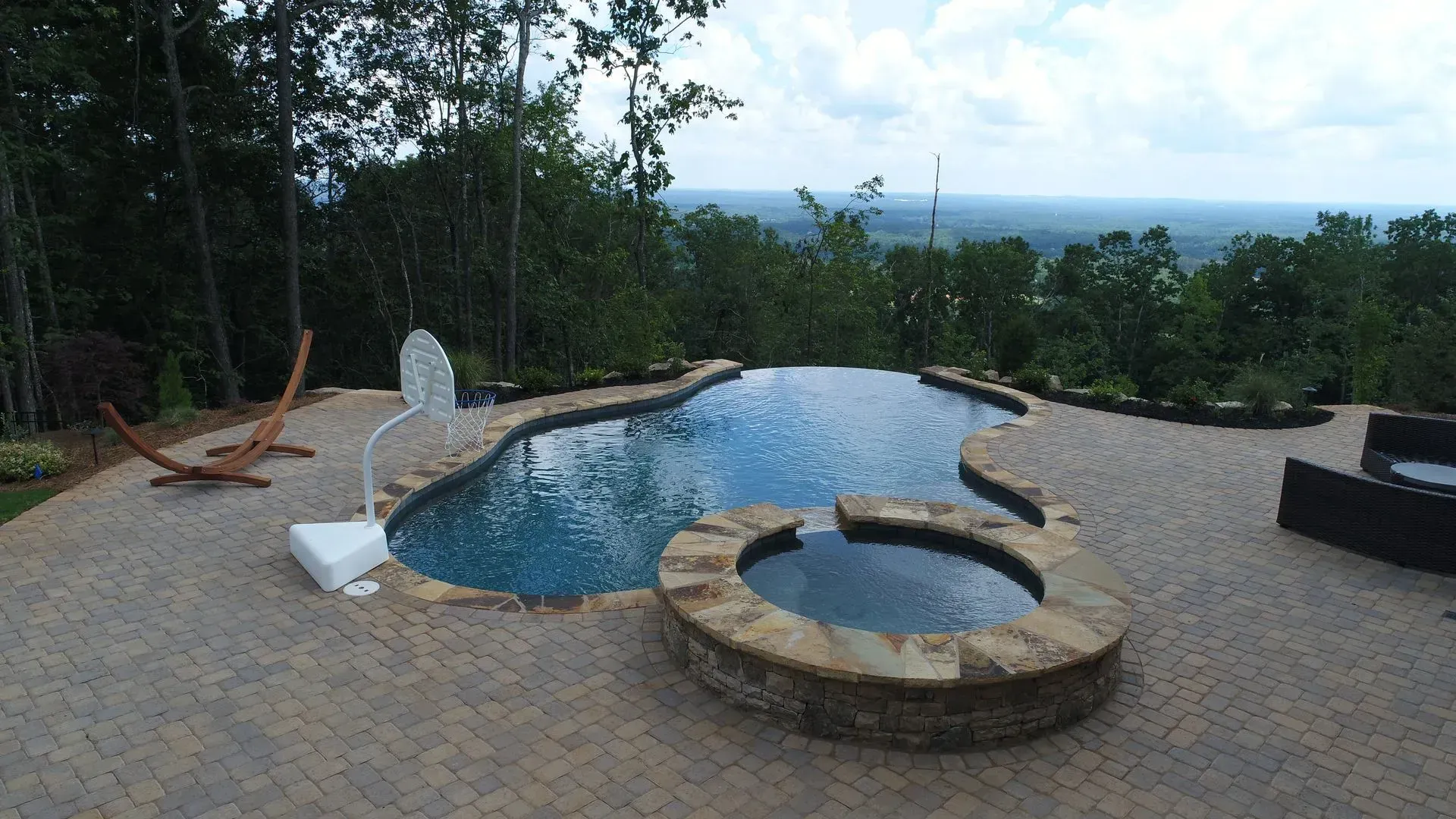 Pool with a spa, overlooking a scenic view with trees, surrounded by stone patio.