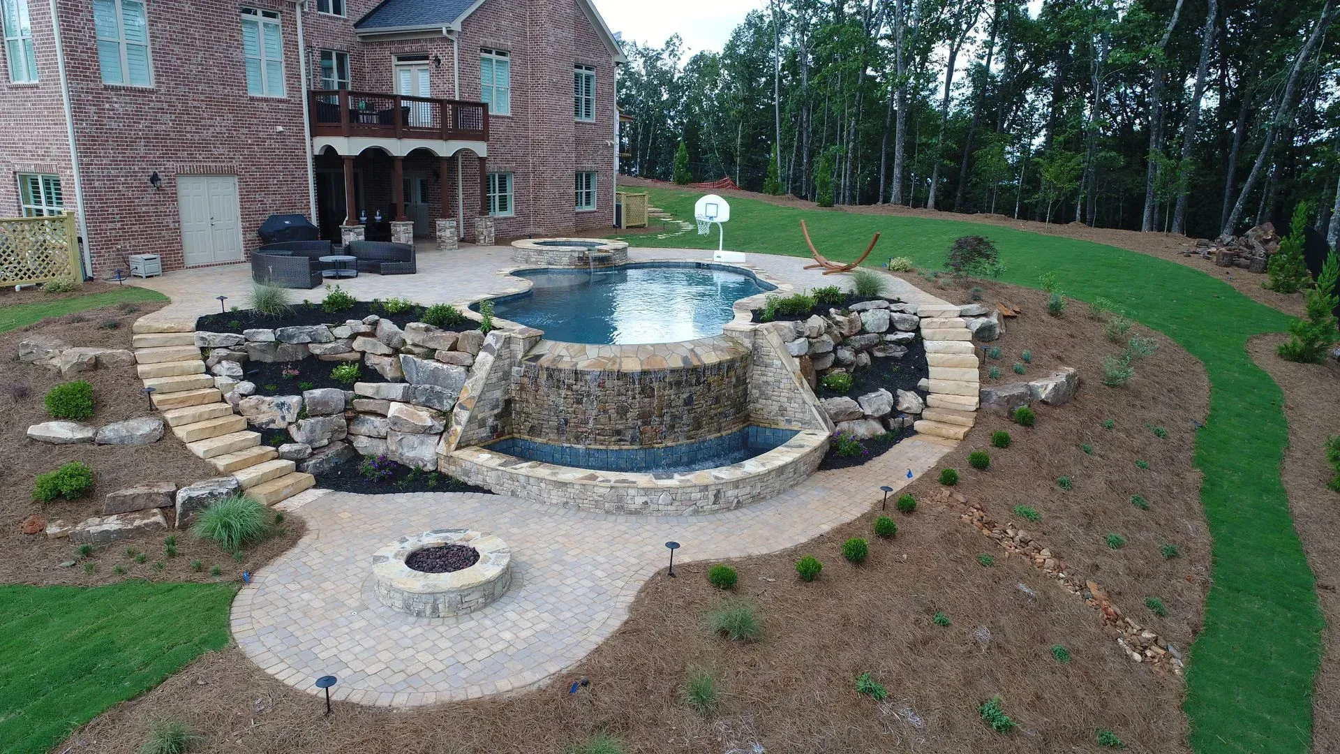 Backyard with pool, waterfall, fire pit, and stone patio, adjacent to a large brick house.