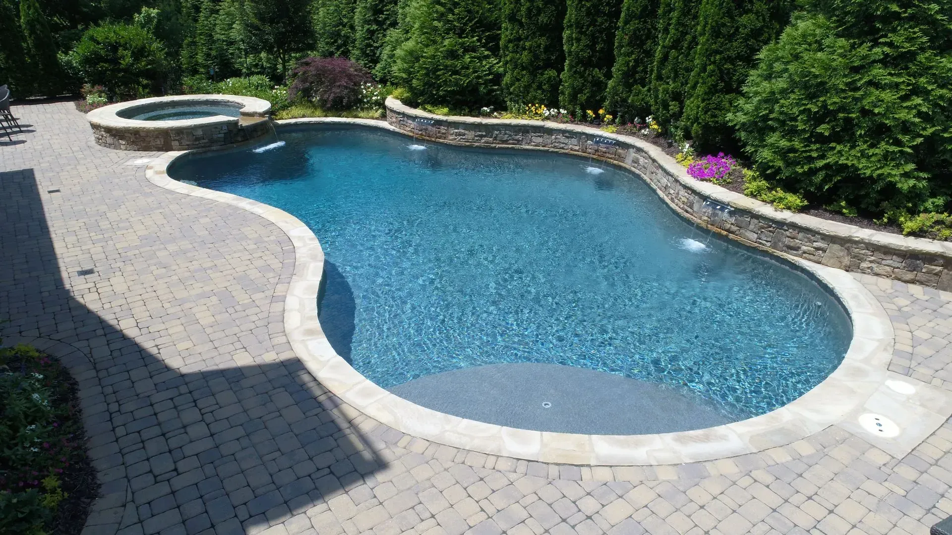 Curving blue swimming pool with attached hot tub, surrounded by stone patio and landscaping.