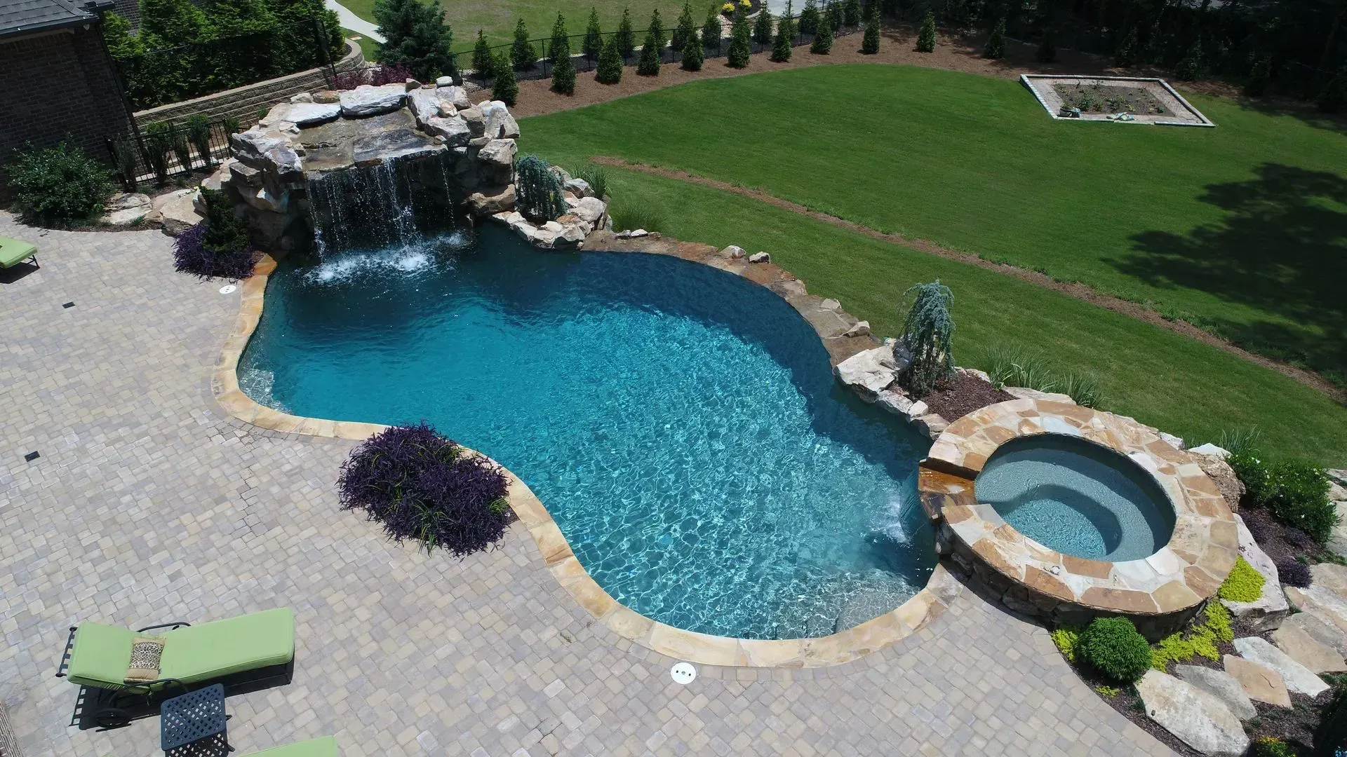 Pool with waterfall, spa, and landscaping in a backyard. Turquoise water, stone accents, green lawn.
