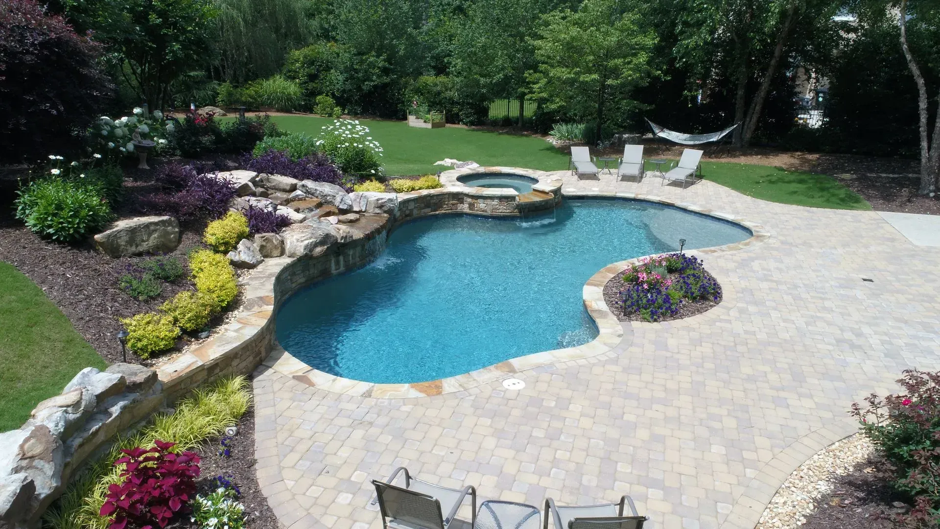 Swimming pool with waterfall feature, surrounded by landscaping, patio, and chairs.