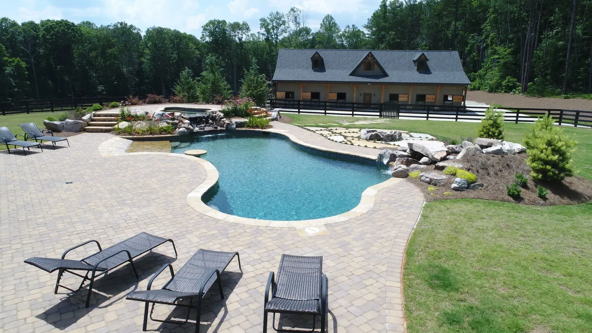 Swimming pool and patio with lounge chairs, next to a barn-like structure and a grassy lawn.