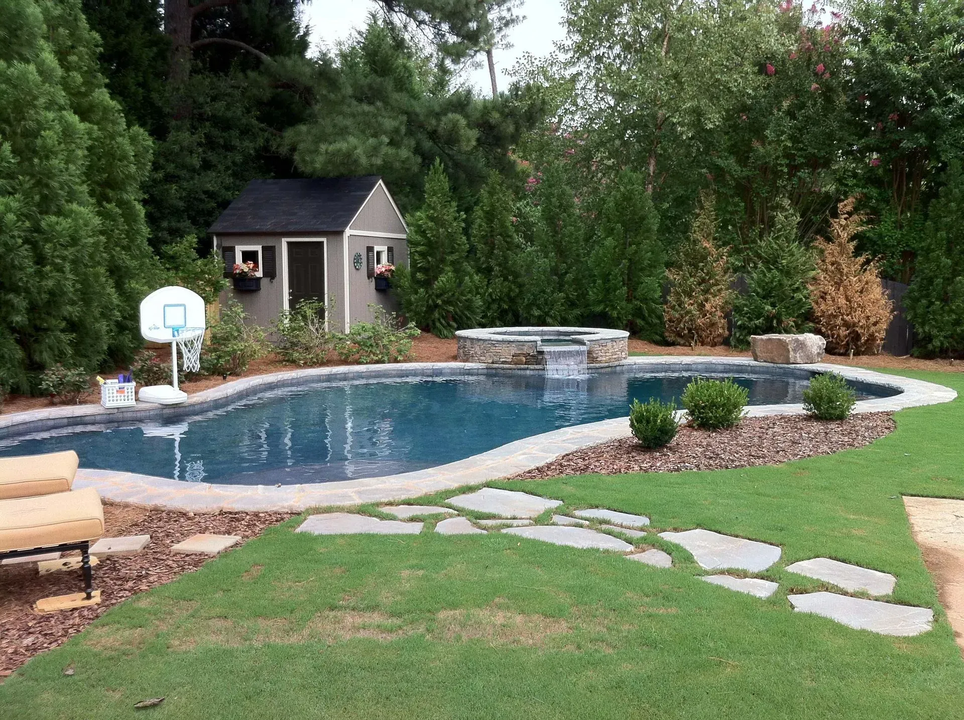 Pool with spa, shed, basketball hoop, and stepping stones in a grassy backyard, surrounded by trees.