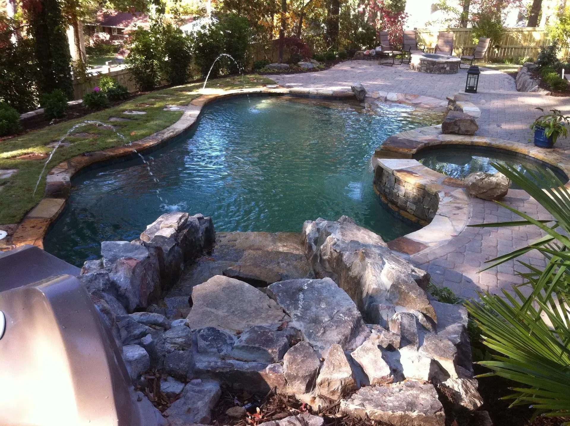 Swimming pool with rocky waterfall, hot tub, and patio area. Green trees and blue water.