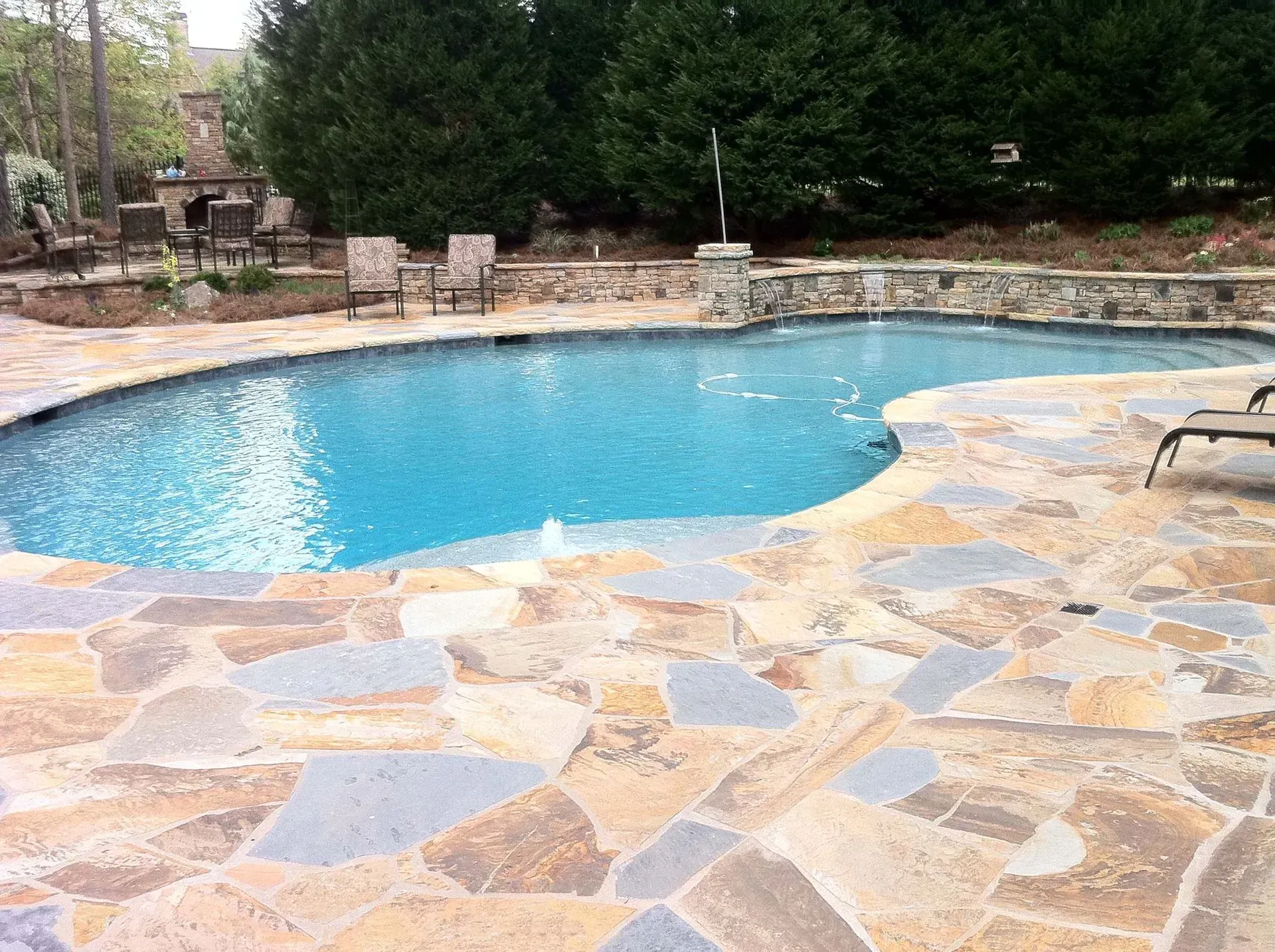 Swimming pool with stone patio. Pool has blue water, surrounded by flagstone. Trees and building in background.