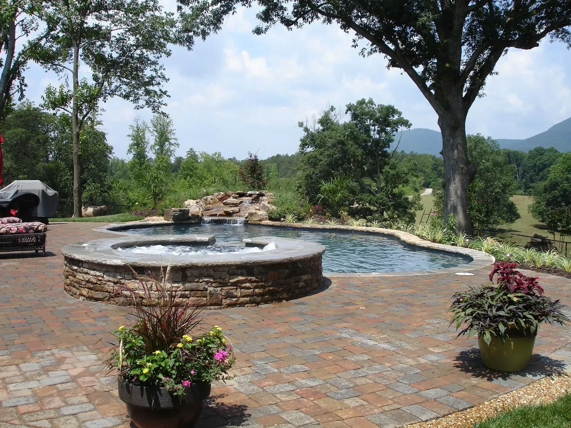 Brick patio with pool, hot tub, and lush landscaping, set against a mountain backdrop.