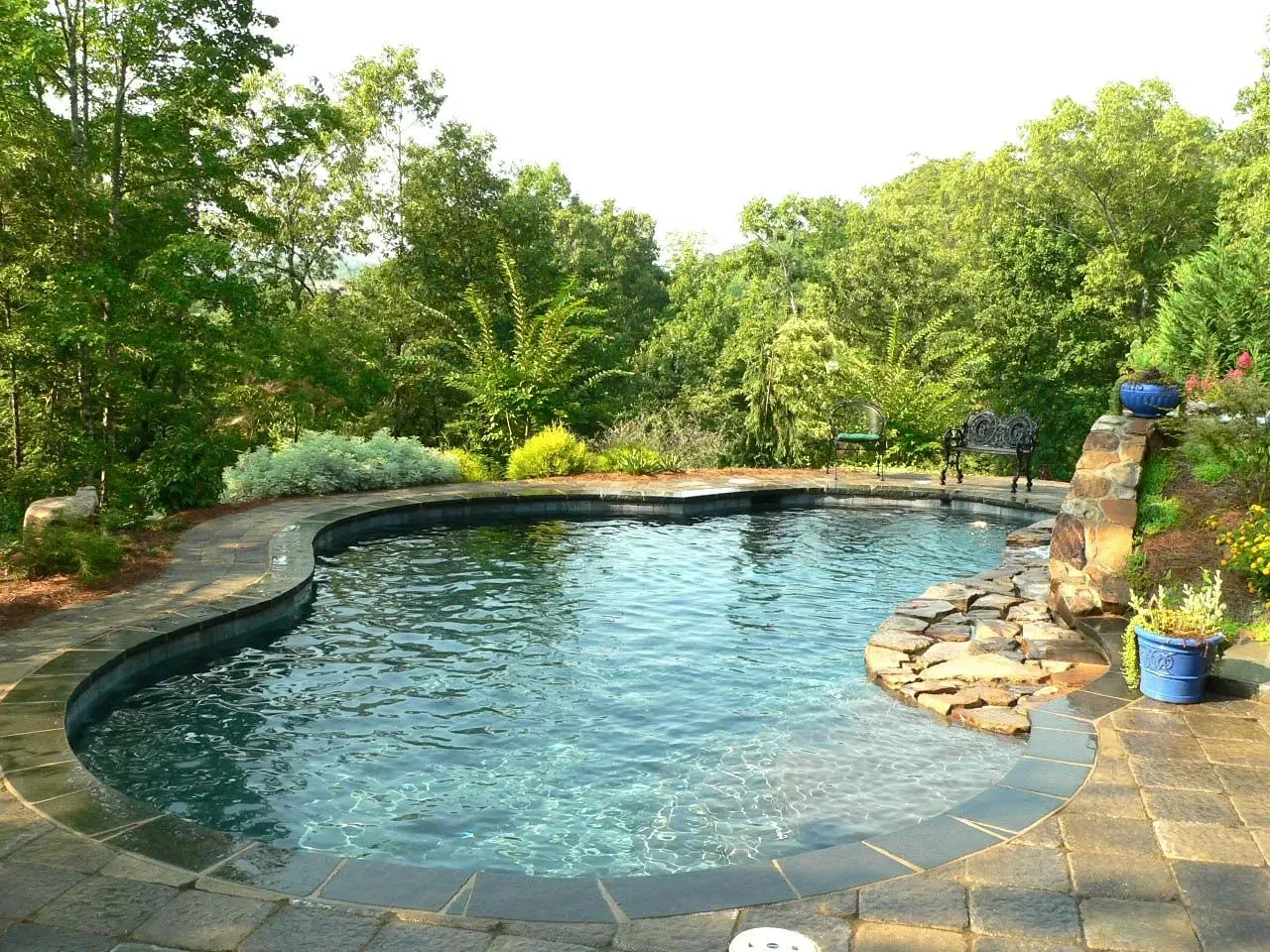 Pool surrounded by stone patio with waterfall and lush greenery in the background.