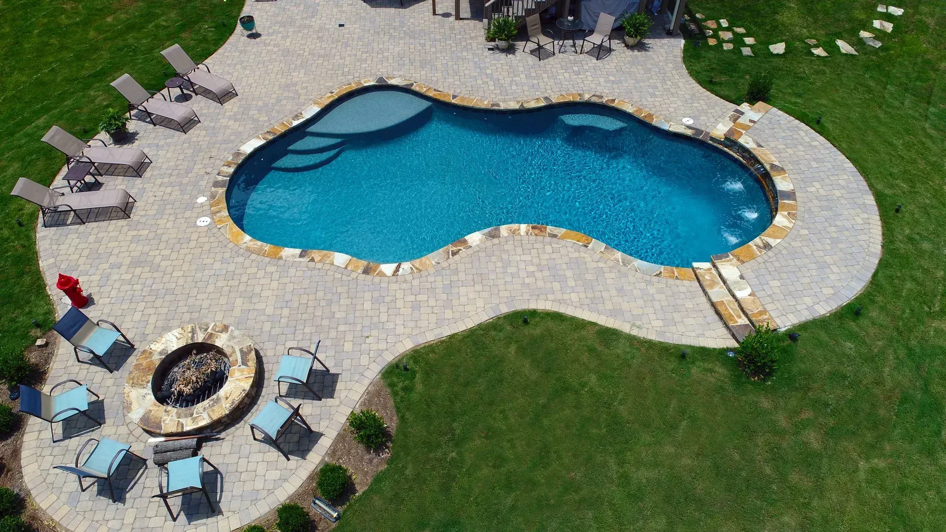 Overhead view of a swimming pool with a stone patio, lounge chairs, and a fire pit in a grassy yard.
