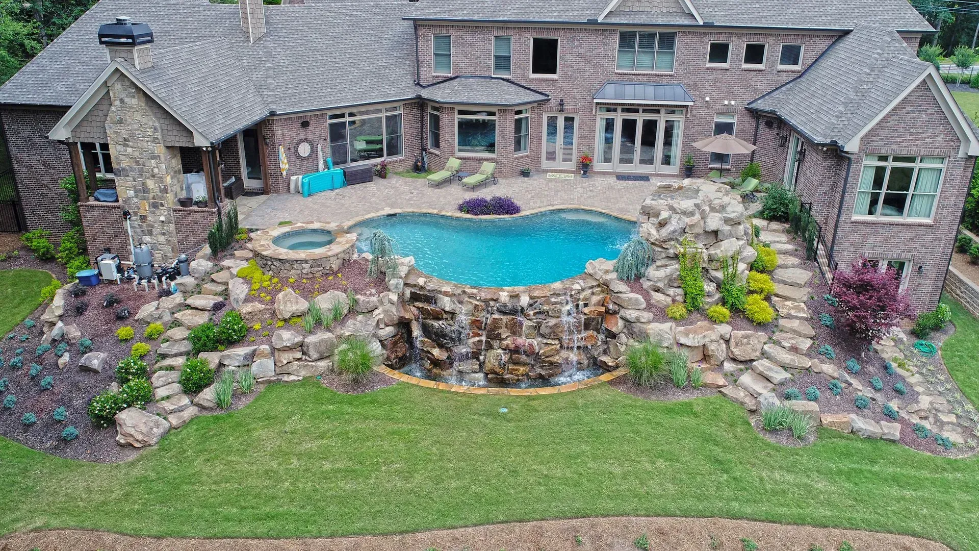 Backyard with a heart-shaped pool and waterfall, surrounded by landscaping, stone steps, and a large brick house.