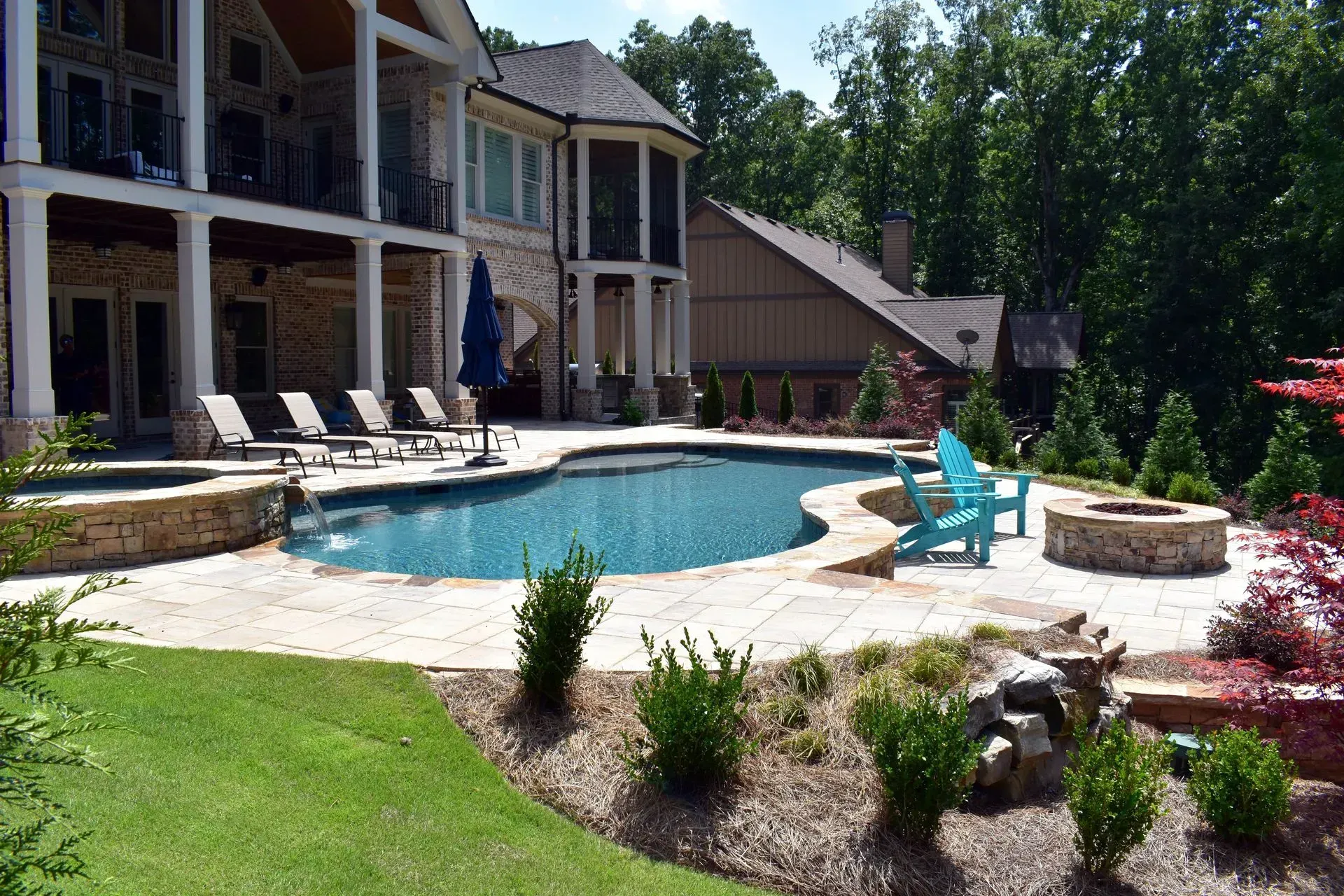 Backyard with pool, hot tub, and fire pit next to a large house with a balcony and landscaping on a sunny day.