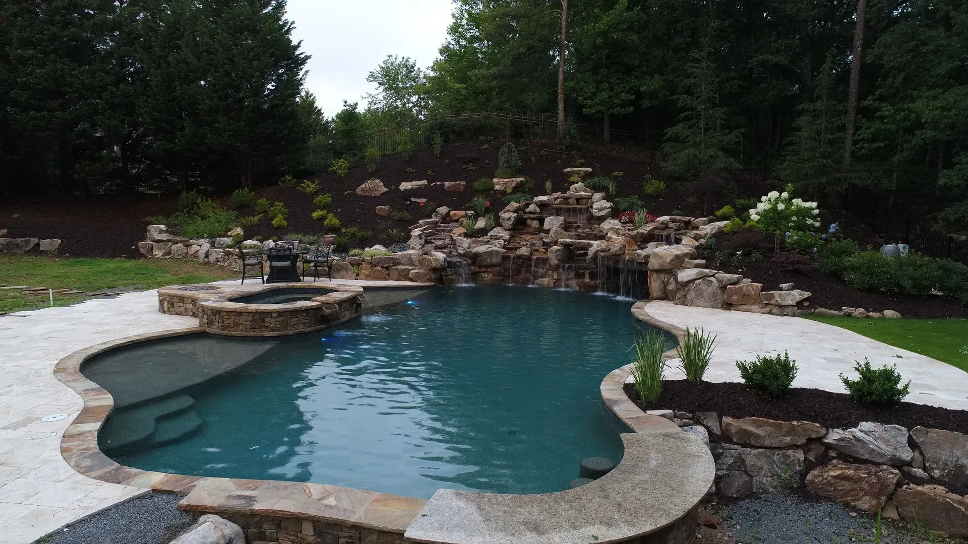 A scenic backyard pool with a waterfall, hot tub, and stone edging surrounded by green grass and trees.