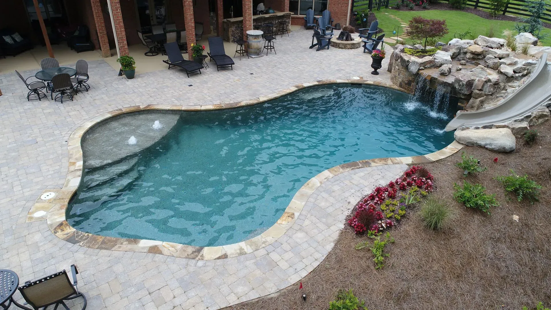 A freeform swimming pool with a waterfall and slide, surrounded by a stone patio and landscaping.