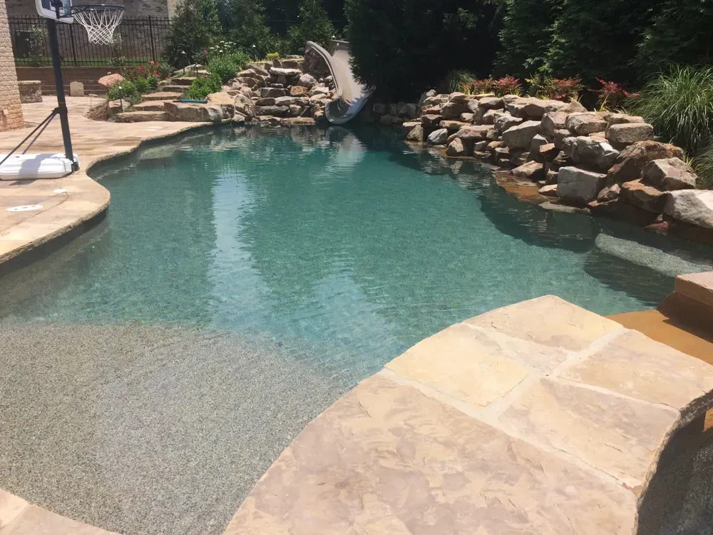 Pool with rock waterfall, slide, and basketball hoop, surrounded by stone patio and landscaping.
