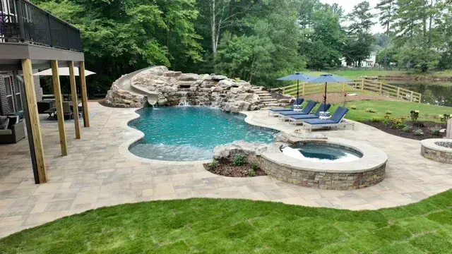 Pool with a waterfall, hot tub, lounge chairs, and an umbrella, surrounded by landscaping and a lake.